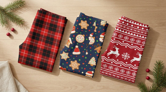 Three pairs of FIERCEPULSE Christmas leggings folded flat on an oak table: red tartan plaid, navy gingerbread cookie print, and crimson Nordic reindeer Fair Isle, styled with pine sprigs and red berries