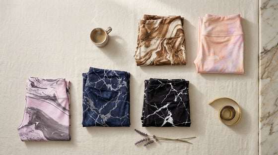 Overhead flat lay of five folded marble-print yoga leggings on cream linen with lavender, ceramic coffee cup, and brushed brass measuring tape