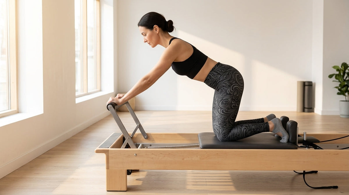 Woman in charcoal 3D Art Deco Yoga Leggings kneeling on a Pilates reformer carriage in a sunlit studio, hands on the foot bar