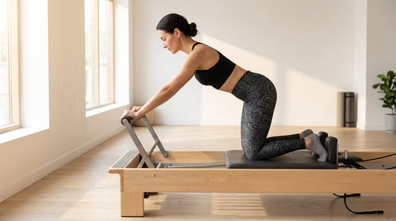 Woman in charcoal 3D Art Deco Yoga Leggings kneeling on a Pilates reformer carriage in a sunlit studio, hands on the foot bar